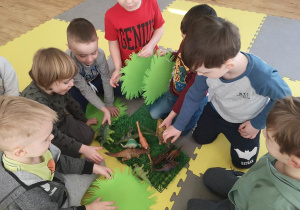 Widok na grupę chłopców, którzy układają prehistoryczny las wykorzystując plastikowe dinozaury, zielone serwetki, trawki.